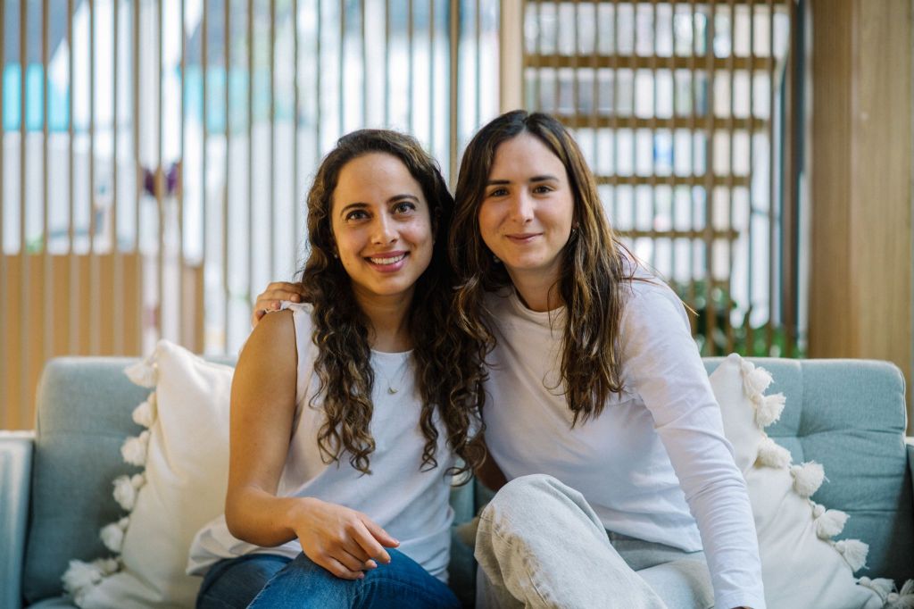 Lorena Ostos and Gio Abramo, co-founders of Plenna, sitting together and smiling, wearing casual white tops, in a warmly lit modern space.